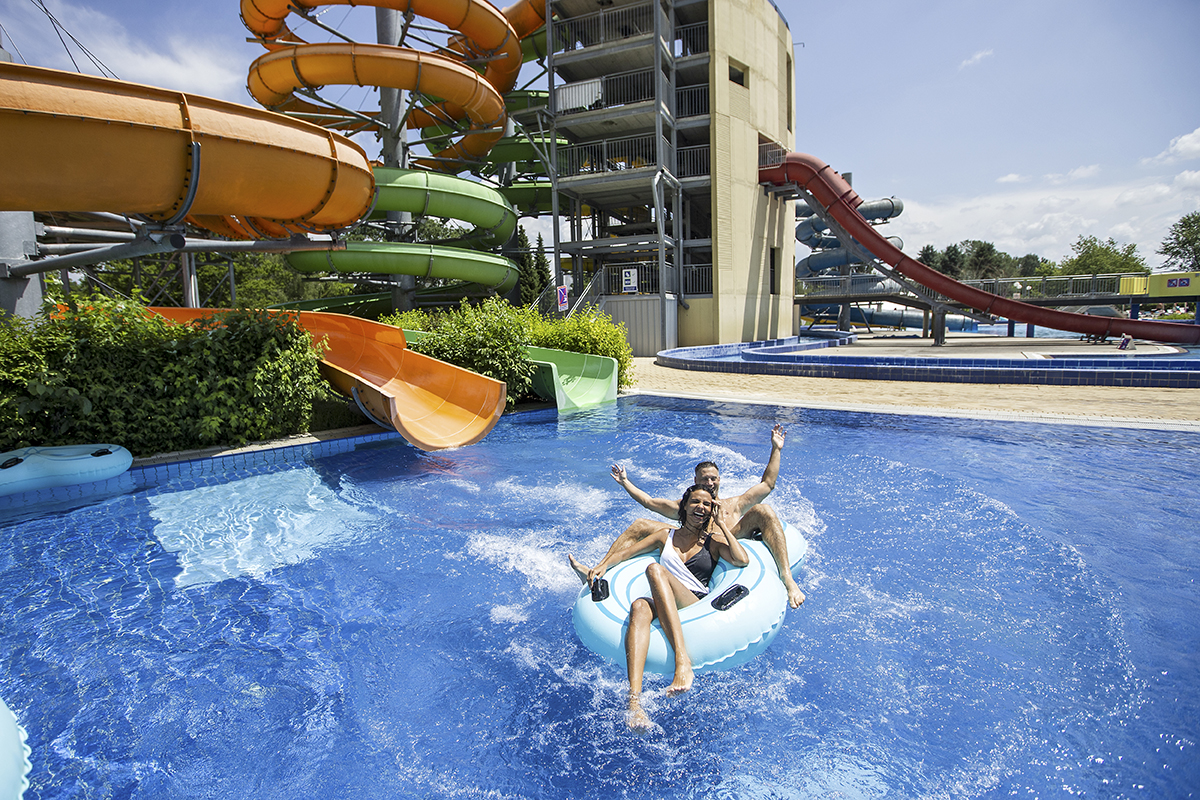 Mlad par se na modri blazini spušča iz oranžnega tobogana v zunanji bazen