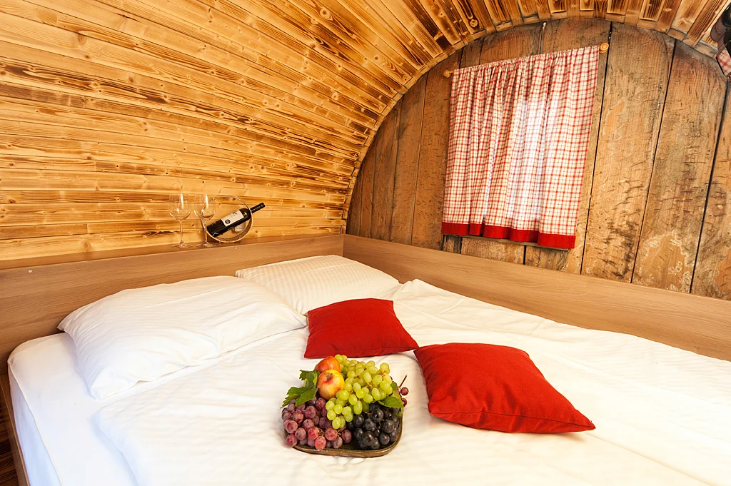 Bed with white linen, red cushions and flowers inside a wooden wine barrel.