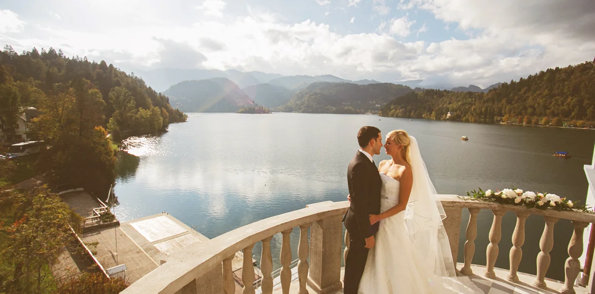 Novoporočenca se v poročni obleki objemata in zaljubljeno gledata na terasi ob jezeru