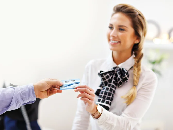 Ženska v beli srajci drži v roki kartico z napisom "ambasador" in jo daje v roke gostu