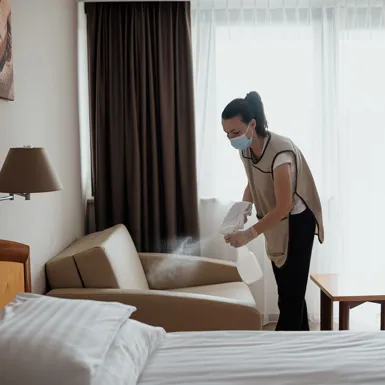 Čistilka v hotelu z zaščitno masko na obrazu dezinficira posteljo v hotelski sobi