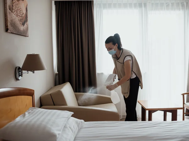 Čistilka v hotelu z zaščitno masko na obrazu dezinficira posteljo v hotelski sobi
