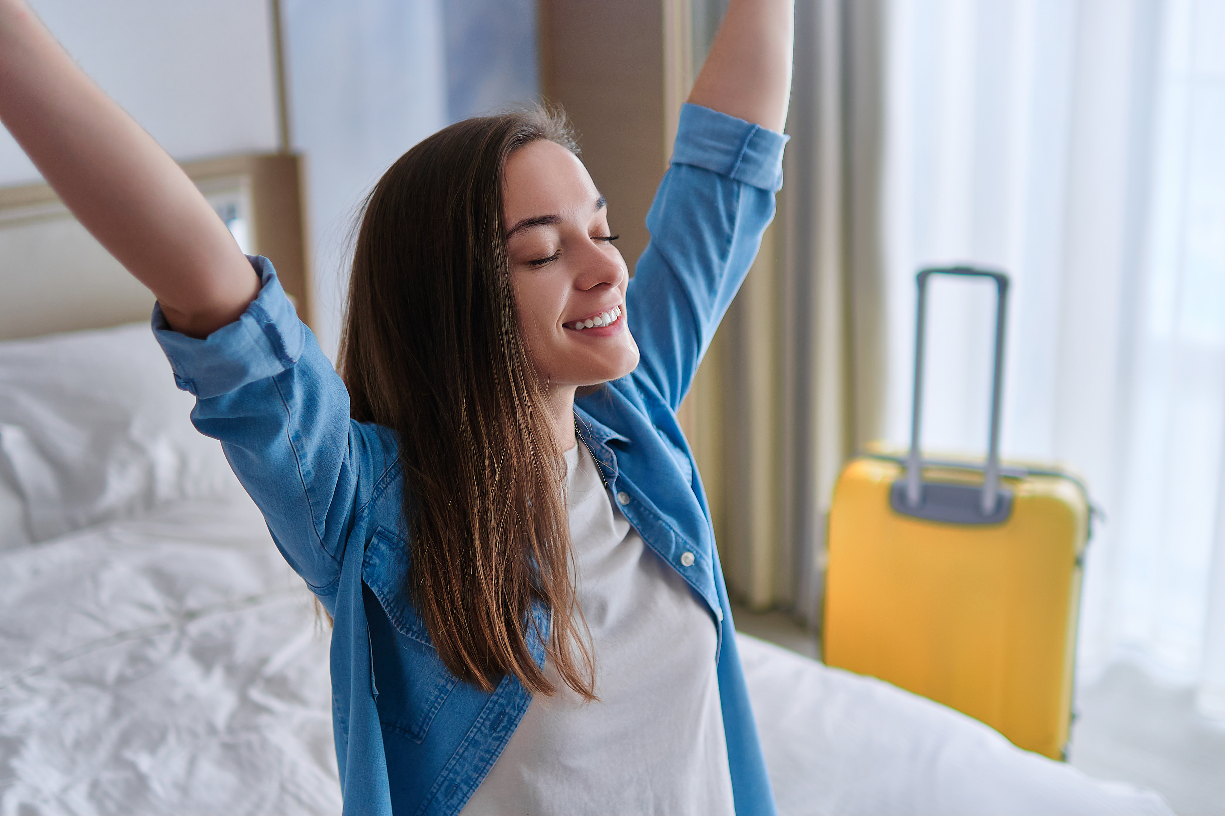 Mlada ženska v hotelski sobi sedi na postelji s kovčkom oknu in dviga roke v zrak