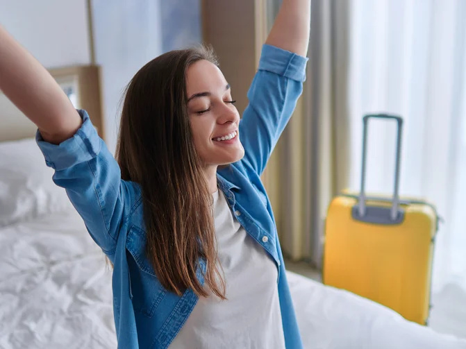Mlada ženska v hotelski sobi sedi na postelji s kovčkom oknu in dviga roke v zrak
