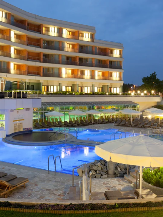 Pogled na osvetljen zunanji bazen, bele dežnike, ležalnike, teraso in hotel