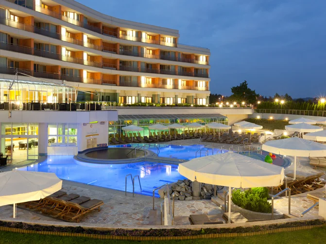 Pogled na osvetljen zunanji bazen, bele dežnike, ležalnike, teraso in hotel